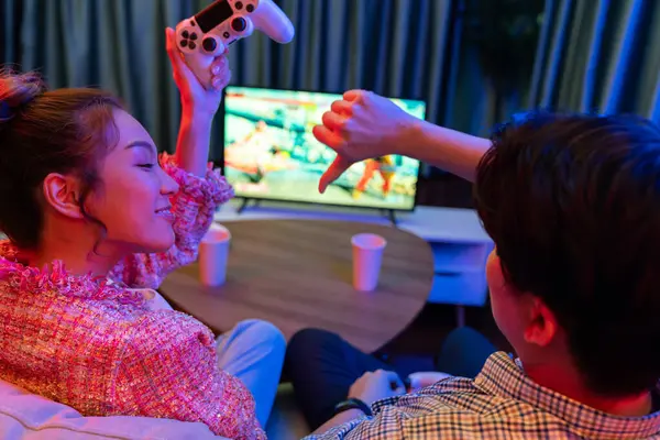 Joystickli birkaç oyuncu dövüş oyunu oynuyor, kadın oyuncu yarışması derecesi arka tarafta TV ekranında modern rahat evinde neon ışıklı oturma odasındaki koltukta oturuyor. İnfobahn.