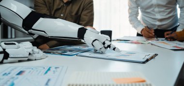 İnsan iş arkadaşlarıyla ortak bir toplantıya katılan bir robot kolu modern ofis masasındaki çizelge ve belgelere işaret ederek, takım çalışması sergiliyor.