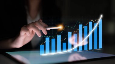 Bir iş kadını, modern analitik ve büyümeyi resmeden çağdaş bir ofiste yükselen grafik ve grafikleri gösteren bir tablet üzerinde mali verilerle uğraşıyor. Etki
