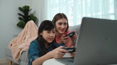 Annem dizüstü bilgisayar kullanarak kanepeye uzanırken, mutlu kız oyun oynuyor ve oturuyor. Beyaz bir ebeveyn ve çekici bir kız, elinde joystick ile birlikte vakit geçirir ve maç kazanmaya odaklanırlar. Pedagoji.