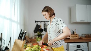 Enerjik anne, mutfak malzemeleri ile çevrili sebzeleri doğrarken modern mutfakta kahvaltı için salata yapıyor. Akıllı kadın temizlikçi yemek pişirir ve yemek hazırlar. Sağlıklı bir yaşam tarzı. Pedagoji.