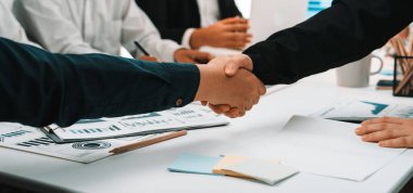 Parlak bir ofis ortamında profesyonellerin el sıkışması, ortaklığı, işbirliğini ve iş dünyasındaki başarılı müzakereleri sembolize eder. SACTR