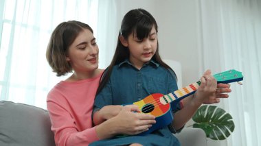 Kafkasyalı bir anne evde akustik müzik öğretirken ukulele çalan mutlu kız. Sevimli çocuk enstrüman öğreniyor. Kafkasyalı mutlu anne ve kız birlikte vakit geçiriyorlar. Pedagoji.