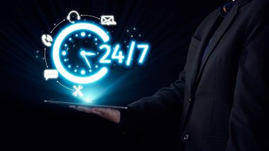 Bir tablet tutan profesyonel bir kişi, modern karanlık bir ortamda bir saat ve iletişim ikonlarının yer aldığı 24 saatlik parlak hizmet uygunluğu kavramını sergilemektedir. Dörtgen