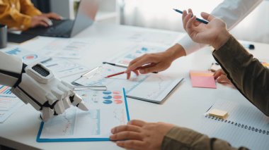 İş haritaları üzerinde insan ve robotik elleri birlikte çalışırken gösteren dinamik bir sahne, iş yerindeki teknoloji ve insan yaratıcılığının birleşimine örnek olarak. Noogenesis