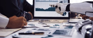 Profesyonel bir toplantı sırasında robot bir elin veri analiz çizelgelerine işaret ettiği modern bir çalışma alanı. Sahne, iş dünyasındaki yenilik ve işbirliğini simgeliyor. Noogenesis.