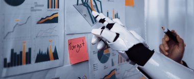 Bir robot el, modern ofiste detaylı iş haritaları ve grafikleri analiz ederken görülüyor. Veri görselleştirme ve teknoloji entegrasyonu sergileniyor. Noogenesis