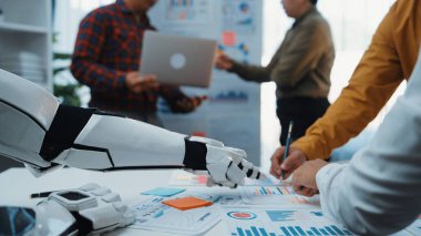 İşbirlikçi bir ortamda dokümanlar, dizüstü bilgisayarlar ve veri analizlerini tartışan profesyonellerle uğraşan bir robot asistanını gösteren modern bir ofis sahnesi. Noogenesis