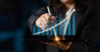 Bir iş adamı dijital bir kalem kullanarak, canlı grafikler ve finansal büyüme ve stratejiyi temsil eden veri eğilimleri sergileyen bir tabletle etkileşime girer. Etki