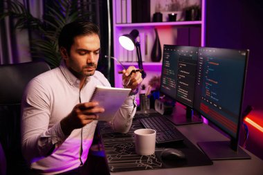 Akıllı bilişim geliştiricisi bilgisayar ekranında yazılım geliştirme kodlaması üzerinde çalışıyor ve en yeni uygulama projesinin notlarını okuyor, program güncellemesi düşünüyor neon ışıklı siber ofiste online web sitesi verileri. Tahmin et..