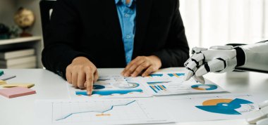 Bir işadamı, modern bir ofiste çalışan bir robotun yardımıyla finansal raporları analiz etmeye odaklanır. Teknolojinin iş dünyasına entegrasyonunu vurgular.