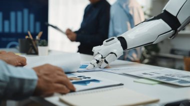 Gelecekteki bir ofis sahnesinde insan eliyle doküman ve grafikler üzerinde uğraşan, dijital bir çalışma alanındaki verimlilik ve işbirliğini vurgulayan robotik bir kol sergileniyor. Noogenesis