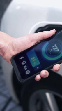 Akıllı telefon akü durumu arayüzünü elinde tutan iş kadını akıllı EV mobil uygulama ile otomobil parkındaki şarj istasyonundan elektrik şarj eden çevre dostu EV araba. İnceleyin