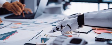 Robot bir kol, modern bir ofiste, teknolojinin ve takım çalışmasının geleceğini gösteren çeşitli grafik ve verileri analiz ederek bir insanla iş birliği yapıyor.