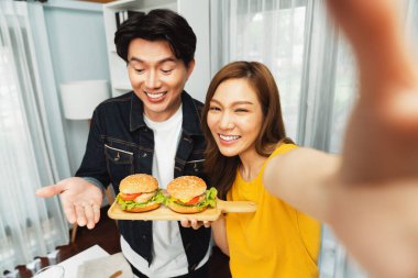 Gülümseyen sunucu şefler akıllı telefonlarıyla fotoğraf çekiyorlar. Özçekim yoluyla. Yemek programında çift hamburger tutuyorlar. İnfobahn.