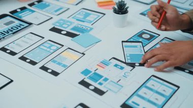 Kağıt prototipleri ve dijital platformlarla kullanıcı arayüzü tasarımı üzerinde çalışan, mobil uygulama geliştirme süreçlerinde takım çalışması ve yaratıcılığı sergileyen ellerin tutulduğu sahne. SACTR