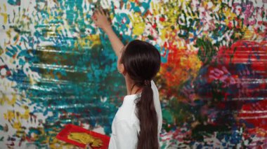 Genç ve güzel bir kızın arka görünüşü renkli duvarı boyamak için elini kaldırır. Çekici bir lise öğrencisi renk paletini tutarken duvarı el iziyle boyuyor. Yaratıcı aktivite. Öğretim