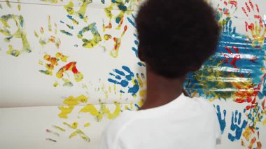Mutlu Afrikalı çocuğun arkadan bakışı, resim dersinde günlük beyaz gömlek giyerken boyalı duvarı renkli eliyle boyadı. Mart öğrencisi yaratıcı sanat çalışması yapmak için el izi kullanıyor. Eğitim. Öğretim