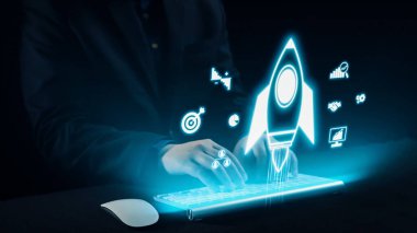 Bir işadamı, parlak bir klavyenin üzerinde, yenilik, dijital teknoloji ve modern bir çalışma alanında başarı hırsını temsil eden holografik bir roket sembolü ile meşgul olur. Dörtgen