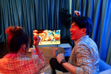 joystickli bir çift, TV ekranında dövüş oyunu oynuyor, erkek, kız arkadaşıyla yarışma seviyesini geri kazanıyor, modern evdeki neon ışıklı oturma odasında kanepeye oturuyor. İnfobahn.