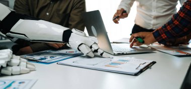 Bir insan ve robot el ofis ortamında birlikte çalışır, teknoloji ve görsel verilerle etkileşim içinde, iş dünyasındaki işbirliğini ve yeniliği sembolize eder.