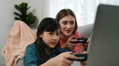 Annem dizüstü bilgisayar kullanarak kanepeye uzanırken, mutlu kız oyun oynuyor ve oturuyor. Beyaz bir ebeveyn ve çekici bir kız, elinde joystick ile birlikte vakit geçirir ve maç kazanmaya odaklanırlar. Pedagoji.