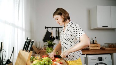 Enerjik anne, mutfak malzemeleri ile çevrili sebzeleri doğrarken modern mutfakta kahvaltı için salata yapıyor. Akıllı kadın temizlikçi yemek pişirir ve yemek hazırlar. Sağlıklı bir yaşam tarzı. Pedagoji.