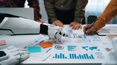 Robotik bir el, verileri analiz eden ve grafikler ve grafiklerle stratejiler geliştiren, modern ofislerde teknoloji ve takım çalışmasının karışımını gösteren bir iş ekibine yardım eder. Noogenesis