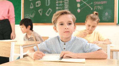 Çekici beyaz çocuk okulda matematik okurken kameraya bakıyor. Çeşitli şirin öğrenciler test yaparken öğretmen Asyalı çocuklara soru sorup tahtaya kağıt doğrultuyor. Pedagoji.