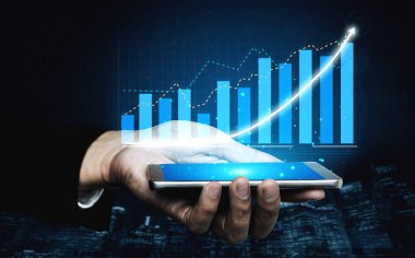 Mavi çizgili ve eğilimli finansal büyüme grafiğini gösteren akıllı telefonu tutan bir elin yakın plan görüntüsü. Arka planda karanlık bir şehir manzarası var. Etki