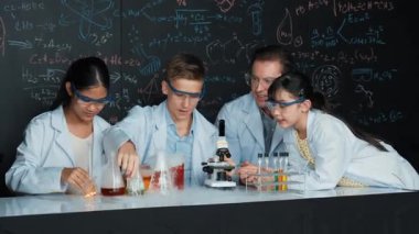 Çok kültürlü bir öğrenci mikroskop altında incelerken güzel bir kız da numuneyi inceliyor. BTMM sınıfında karatahta bilim teorisi deneyi yapan liseli çocuklara bakan öğretmen. Öğretim