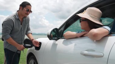 İlerici mutlu aile kavramı elektrikli araçla rüzgar çiftliğinde eğleniyor. Rüzgar türbini jeneratöründen gelen temiz yenilenebilir enerji ile şarj edilen elektrikli araç..