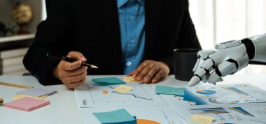 Bir iş adamı, veri tabloları ve grafiklerini analiz ederken robotik bir kol ile işbirliği yapar, modern çalışma ortamlarında teknolojinin entegrasyonunu vurgular. Noogenesis