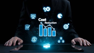 Bir işadamı, gösterişli ve karanlık bir ortamda eğilimleri ve finansmanı temsil eden grafikler ve simgelerin yer aldığı maliyet düşüşüne odaklı dijital analizler sergiliyor. Dörtgen
