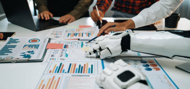 İş ortamlarında yenilik ve üretkenliği vurgulayan veri analiz grafikleri ve araçların yer aldığı modern bir çalışma alanı, insanlar ve robotlar arasındaki işbirliğini sergiliyor.