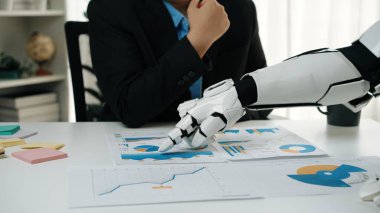 Takım elbiseli bir insan robot kolla birlikte iş verilerini de analiz eder. Bu resim, teknoloji ve insan uzmanlığının karar verme mekanizmasında bütünleşmesini sembolize ediyor.