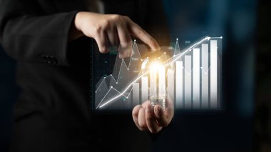 Bir iş adamı parlak fikirleri sembolize eden parlayan bir ampulü işaret eder. Arkasında, yukarı bakan grafikler kurumsal bir ortamda finansal büyüme ve analizi gösteriyor. Etki