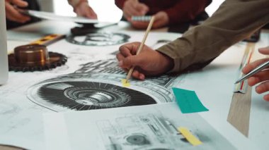 Bir grup mühendis, bir atölyede takım çalışması ve tasarım hassasiyetini vurgulayarak ayrıntılı teknik çizimler ve makine bileşenlerini işbirliği içinde analiz ediyor. SACTR