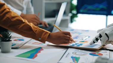 Bir robot asistan analitik çalışmalarını desteklerken iş adamları performans çizelgeleri üzerinde işbirliği yapıyorlar. Bu sahne yenilik ve takım çalışmasını modern ofis ortamında sergiliyor. Yaramazlık