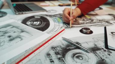 Yaratıcı araçlar ve teknolojiyle dolu modern bir çalışma alanında detaylı planlar ve mekanik diyagramlar üzerinde çalışan tasarımcıların yakın çekim görüntüsü. SACTR