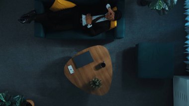 Stresli proje yöneticisinin telefon kullanarak akıllı ekiple konuşmasının en üst görüntüsü. Profesyonel bir iş adamı laptopta çalışırken iş arkadaşını arar ve kanepede yatar. Uzaktan kumanda. Müdürlük.