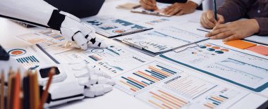 Modern bir çalışma alanı sahnesinde robot ve insan birlikte çalışıyor, çeşitli veri tabloları ve raporlarını analiz ediyor, iş dünyasındaki yenilik ve teknolojiyi somutlaştırıyor. Noogenesis