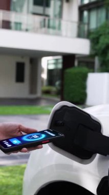 Modern yaşam tarzı bir kadın EV şarj cihazını ev şarj istasyonuna yerleştirir ve akıllı telefondan EV batarya durumunu etkinleştirir. Gelecekçi enerji sürdürülebilirliği ve elektrikli araba. İnceleyin