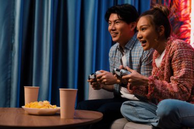 Neşeli, gülümseyen bir çiftin koltuğunda oturup oyun oynuyorlar. Oyun konsolu, hazır aperatifler ve içecekler, modern neon ışıklı, rahat oturma odasında eğlenceli bir rekabet seviyesinde. İnfobahn.