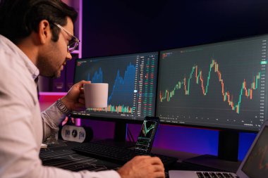 Zeki tüccar işadamı kahve fincanı içiyor, neon ışıklı ofisteki arama pazarında yüksek karlı finans teknolojisiyle bilgisayar üzerinde iki dinamik borsa yatırım ekranını analiz ediyor. Tahmin et..