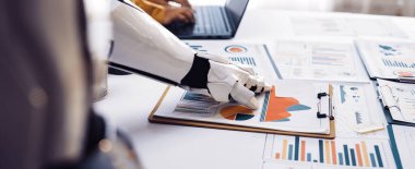 Bir robot el masadaki analiz belgeleriyle etkileşime girerken bir insan bir dizüstü bilgisayar üzerinde çalışır, teknoloji ve veri analizlerinde işbirliği gösterir.