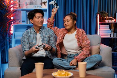 Mutlu çiftin oturduğu kanepe joysticklerle video oyunu oynuyor, meydan okuyan gülümseyen kadın, evdeki modern neon ışıklı oturma odasındaki mücadeleyi kazanmak için yumruk kaldırıyor. İnfobahn.