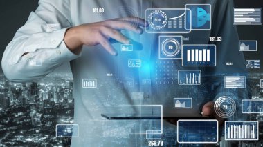 Bir iş adamı, tablet üzerinde veri görselleştirmeleri ve analizlerinin gösterildiği dijital bir arayüzle meşgul olurken, geceleri hareketli bir şehir silüeti sahneyi büyütüyor. Xenic