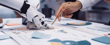 Yenilikçi iş çözümleri için çeşitli veri tabloları ve grafikleri analiz eden, robot el ve insan eli arasındaki işbirliğini gösteren modern bir ofis sahnesi.