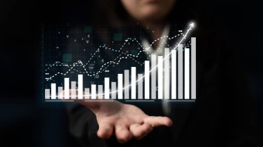 Bir iş adamı, modern bir kurumsal ortamda veri analizlerini gösteren finansal büyüme ve eğilimleri temsil eden dijital grafikler ve grafikler sergiliyor. Etki